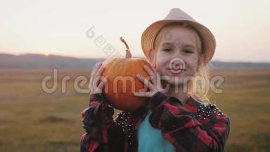年轻的女农民戴着帽子，手里拿着橙色的南瓜，在日落的时候在田野里。 南瓜收获。 感恩节感恩节